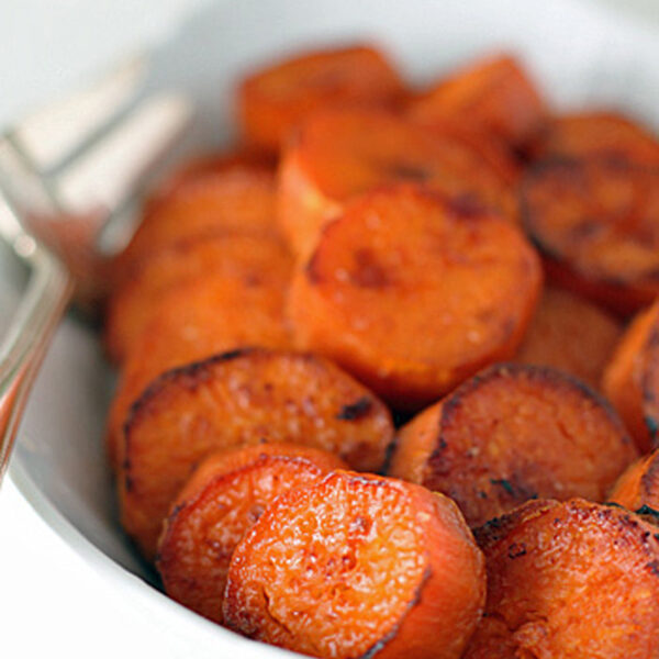Sliced, roasted sweet potatoes with a golden-brown, caramelized surface arranged in a white dish, with a fork partially visible in the background.