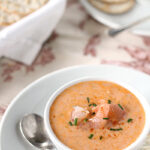 A bowl of creamy shrimp bisque garnished with chives and shrimp sits on a white plate with a spoon and crackers; more crackers are visible in the background.