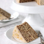 A slice of layered cake with light brown frosting and visible nut pieces sits on a white plate with a fork, with an additional cake slice and cake stand in the background.