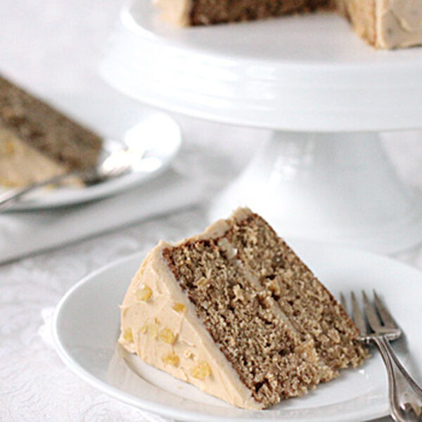 A slice of layered cake with light brown frosting and visible nut pieces sits on a white plate with a fork, with an additional cake slice and cake stand in the background.