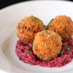 Three golden-brown, crispy arancini balls are served on a bed of vibrant purple beet risotto, garnished with fresh green chives, on a white plate.