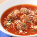 A bowl of tomato-based soup with several meatballs, sprinkled with chopped herbs and crumbled cheese, served on a white plate with a spoon nearby.