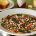 A bowl of soup with black-eyed peas, greens, carrots, and ham pieces sits on a table. In the background are cornbread muffins, a stick of butter, and a bottle of hot sauce.