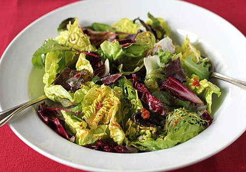 A white bowl filled with a mixed green salad, including leafy greens and radicchio, topped with dressing. A fork rests inside the bowl, which is placed on a red tablecloth.
