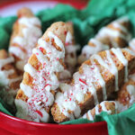 A red bowl lined with green tissue paper holds several biscotti cookies drizzled with white icing and sprinkled with crushed peppermint candy.