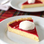 A slice of cheesecake with a layer of red fruit topping and a dollop of whipped cream on a white plate, set on a table with a red plaid tablecloth.