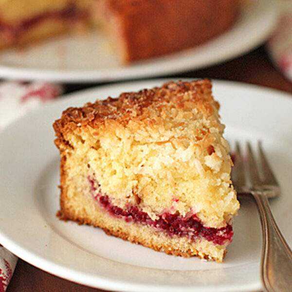 A slice of coffee cake with a layer of raspberry filling and crumbly topping sits on a white plate beside a fork, with a patterned napkin and another plate in the background.