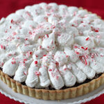 A chocolate pie with a decorative whipped cream topping, sprinkled with crushed peppermint candy, sits on a white plate against a red background.