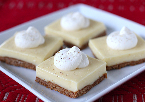 Four square dessert bars with a creamy yellow filling and a brown crumb crust, each topped with a dollop of whipped cream, are arranged on a white plate over a red patterned surface.