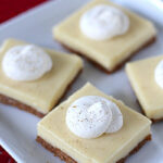 Four square dessert bars with a light cream layer and a brown crust, each topped with a swirl of whipped cream, are arranged on a white plate against a red background.