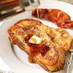 A plate with two slices of French toast topped with butter and syrup being poured, with two strips of crispy bacon on the side and a fork resting on the plate.