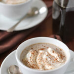 A white cup filled with a creamy hot beverage topped with whipped cream and a sprinkle of cinnamon sits on a saucer with a spoon. Another cup and a glass bottle are blurred in the background.