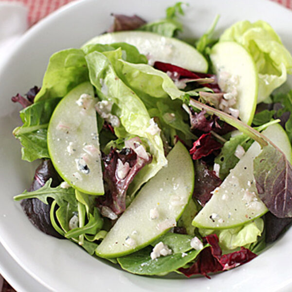 A fresh green salad with mixed lettuce, arugula, and thin apple slices, topped with crumbled cheese on a white plate. A fork and napkin are placed beside the plate.