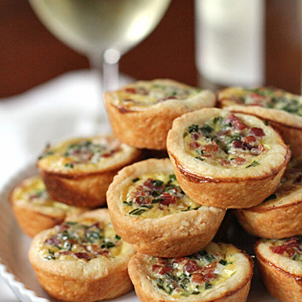 A plate of mini quiches with golden crusts, filled with herbs and diced toppings, is stacked on a white pedestal dish. A glass of white wine sits in the blurred background.