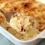 A spoonful of creamy, golden-brown baked casserole is being lifted from a white baking dish with a browned, cheesy topping.