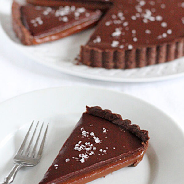 A slice of chocolate caramel tart sprinkled with sea salt sits on a white plate with a fork, while the remaining tart with a missing slice is in the background.