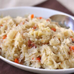 A close-up of a white dish filled with sauerkraut mixed with shredded carrots and small pieces of meat, garnished with black pepper, and a spoon resting on the side.