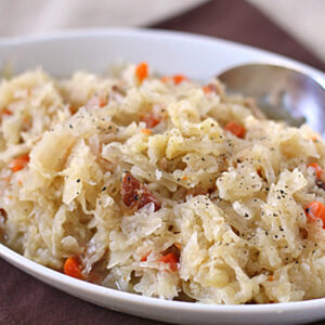 A close-up of a white dish filled with sauerkraut mixed with shredded carrots and small pieces of meat, garnished with black pepper, and a spoon resting on the side.