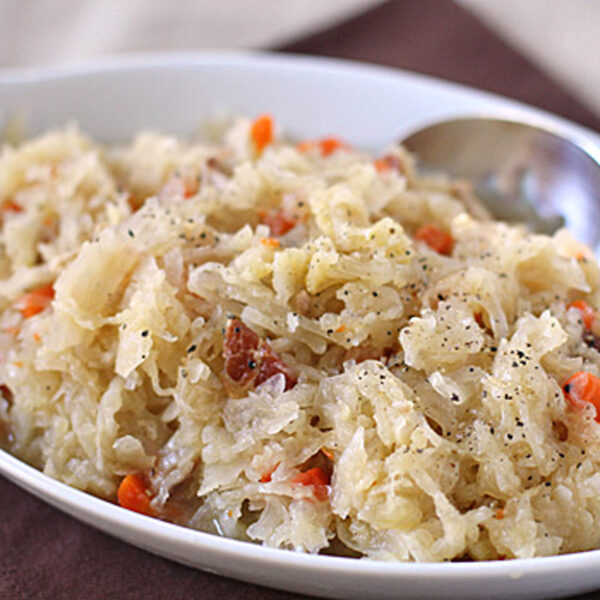 A close-up of a white dish filled with sauerkraut mixed with shredded carrots and small pieces of meat, garnished with black pepper, and a spoon resting on the side.