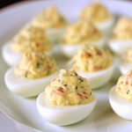 A white plate filled with several deviled eggs, each halved and topped with a creamy, yellow, seasoned filling containing visible herbs.