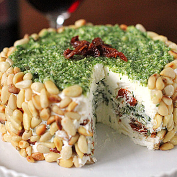 A cheese wheel covered in pine nuts with a green herb topping, garnished with sundried tomatoes. A slice is cut out, revealing layers of cheese, herbs, and sundried tomatoes inside, on a white plate.