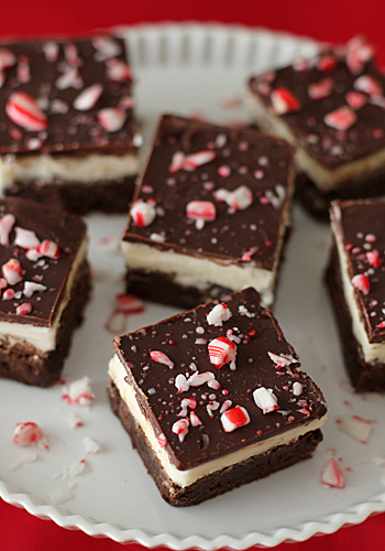 Squares of layered brownies topped with crushed peppermint candy are arranged on a white plate. The brownies have a chocolate topping, a creamy middle layer, and a fudgy base. Red and white candy pieces decorate the top.