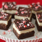 Brownie squares with a white creamy layer and a chocolate topping, sprinkled with crushed peppermint candy, arranged on a white plate against a red background.