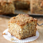 A close-up of a square slice of coffee cake with a crumbly cinnamon streusel topping, placed on a white paper liner. More cake is visible on a cooling rack in the background.
