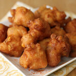 A white plate piled with golden-brown, crispy corn fritters sits on a patterned cloth atop a wooden surface.