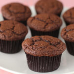 A group of chocolate muffins in dark brown paper wrappers are arranged on a white plate, set against a softly blurred pink background.
