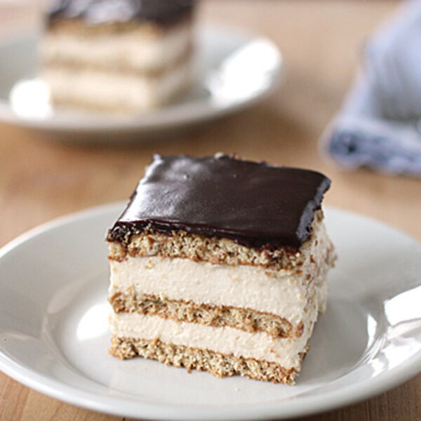 A square slice of layered dessert with cream filling and a glossy chocolate topping is served on a white plate, with another similar dessert and utensils blurred in the background.