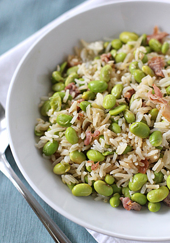 A white bowl filled with rice, edamame beans, and small pieces of bacon, placed on a white napkin with a fork next to it.