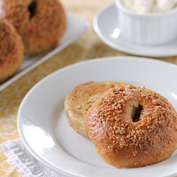 A toasted bagel cut in half sits on a white plate, topped with a crispy, brown coating. In the background, more bagels and a small dish of cream cheese are visible on a patterned yellow tablecloth.
