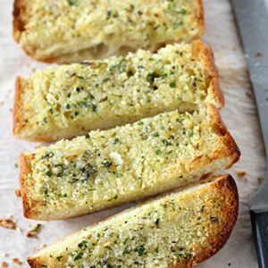 Four slices of golden, crispy garlic bread with chopped herbs and melted butter are arranged on parchment paper next to a large knife.