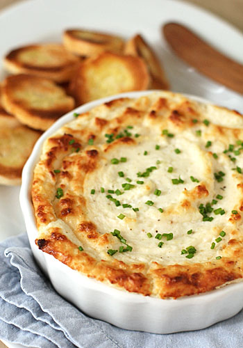 A baked dip topped with chopped chives is served in a round white dish, with toasted baguette slices and a wooden spreader on a white plate in the background. A folded blue napkin is placed underneath the dish.