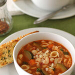 A white bowl of pasta and vegetable soup topped with grated cheese sits on a plate next to a slice of garlic bread. Another bowl of soup and a pepper shaker are visible in the background.