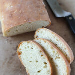 A loaf of rustic bread with a golden crust is partially sliced on a wooden surface, with three slices fanned out in front and a bread knife lying beside it.