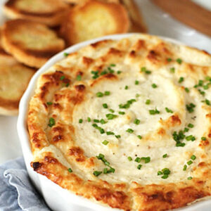 A baked dish of creamy dip topped with chopped chives is served in a white round baking dish, alongside golden toasted baguette slices on a white plate with a wooden spreader.