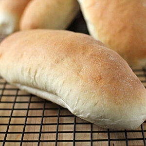 A freshly baked hoagie roll rests on a cooling rack, with other similar rolls in the background. The bread has a light golden-brown crust and a soft texture.