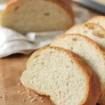 Sliced loaf of white bread on a wooden surface, with crumbs scattered around and a white cloth in the background.