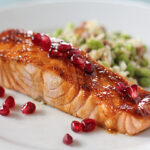 A glazed salmon fillet topped with pomegranate seeds is served on a white plate next to a portion of rice mixed with green vegetables. A fork rests on the side.