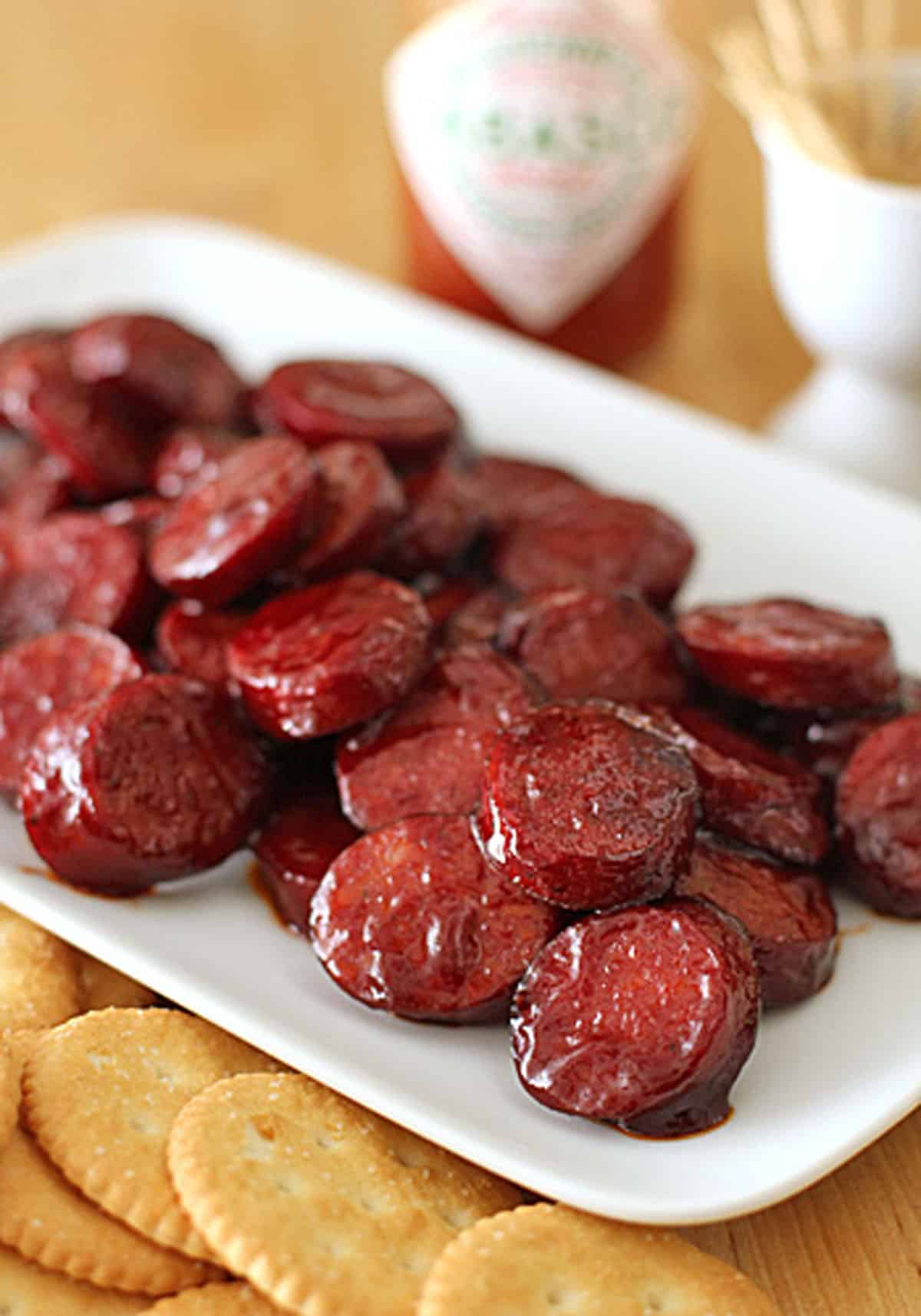 A white plate filled with glazed sausage slices sits next to a stack of round crackers. A bottle of hot sauce and a container with toothpicks are blurred in the background.