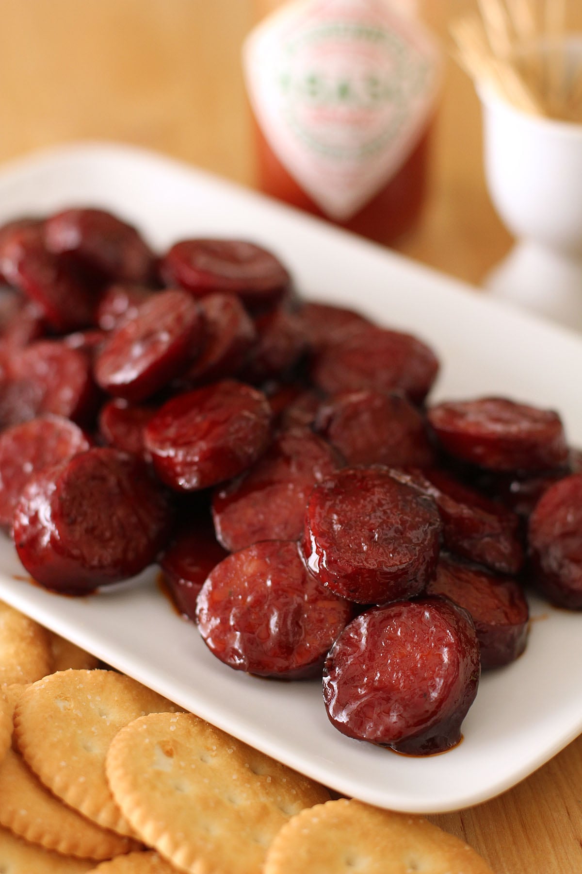 A platter of sliced pomegranate glazed sausage bits served with round crackers and a bottle of hot sauce.