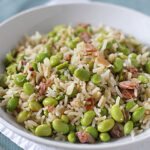 A white bowl filled with rice, edamame beans, and small pieces of meat, placed on a white napkin with a spoon nearby. The dish appears fresh and colorful.