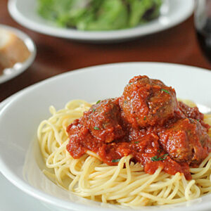 A white bowl filled with spaghetti topped with tomato sauce and meatballs; a plate of bread, a salad, and a glass of red wine are blurred in the background.