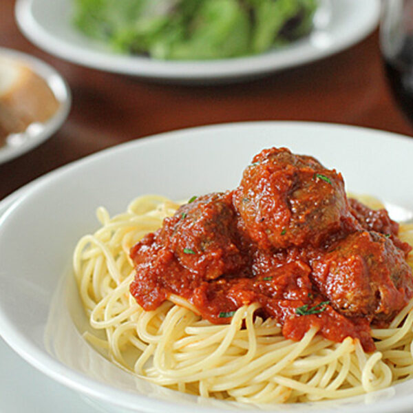 A white bowl filled with spaghetti topped with tomato sauce and meatballs; a plate of bread, a salad, and a glass of red wine are blurred in the background.