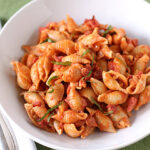 A white bowl filled with shell pasta topped with tomato sauce and garnished with fresh basil, placed on a green tablecloth next to a fork and napkin.