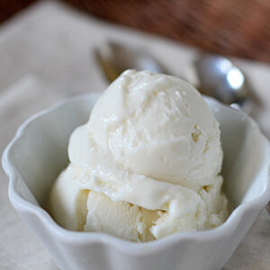 A white bowl filled with two scoops of vanilla ice cream sits on a beige napkin, with two spoons placed in the background.