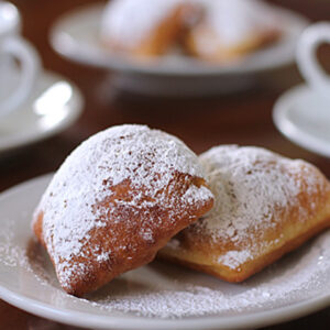 Two beignets dusted with powdered sugar on a white plate, with blurred cups of coffee and more pastries in the background.