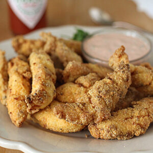 A plate of crispy, golden-brown fried catfish strips is served with a small bowl of creamy dipping sauce on the side, all set on a light-colored plate.